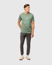 A man wearing a Psycho Bunny MENS MALTON GRAPHIC TEE - B6U795D200 in green, featuring a bunny skull and crossbones logo, paired with gray pants and beige shoes, stands against a plain light gray background. He has his right hand in his pocket and is looking to his left.