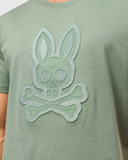 A person wearing the Psycho Bunny MENS MALTON GRAPHIC TEE - B6U795D200, a green Peruvian-crafted t-shirt featuring an embroidered rabbit skull with crossbones design. The graphic includes large circular eyes and two long ears, creating a playful yet slightly edgy look. This comfortable tee is made from soft Pima cotton.