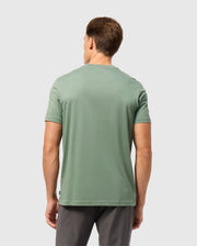 A man with short brown hair stands with his back to the camera, wearing a light green Psycho Bunny MENS MALTON GRAPHIC TEE - B6U795D200 and dark gray pants against a plain light gray background.