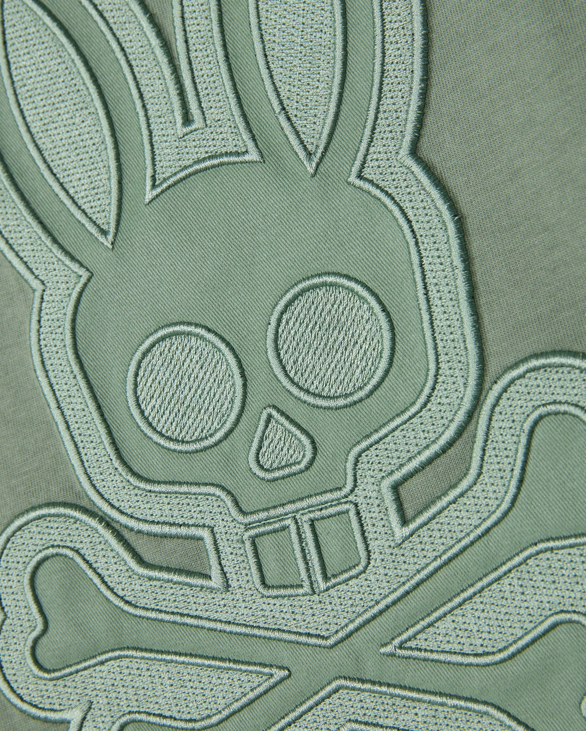A close-up shot of the embroidered patch on the Pima cotton fabric background of the Psycho Bunny MENS MALTON GRAPHIC TEE - B6U795D200, showcasing a bunny skull and crossbones design in light green thread. The intricate stitching lends a three-dimensional feel to the design, echoing Peruvian-crafted artistry.