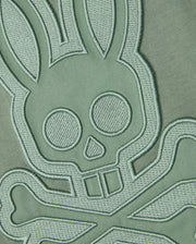 A close-up shot of the embroidered patch on the Pima cotton fabric background of the Psycho Bunny MENS MALTON GRAPHIC TEE - B6U795D200, showcasing a bunny skull and crossbones design in light green thread. The intricate stitching lends a three-dimensional feel to the design, echoing Peruvian-crafted artistry.