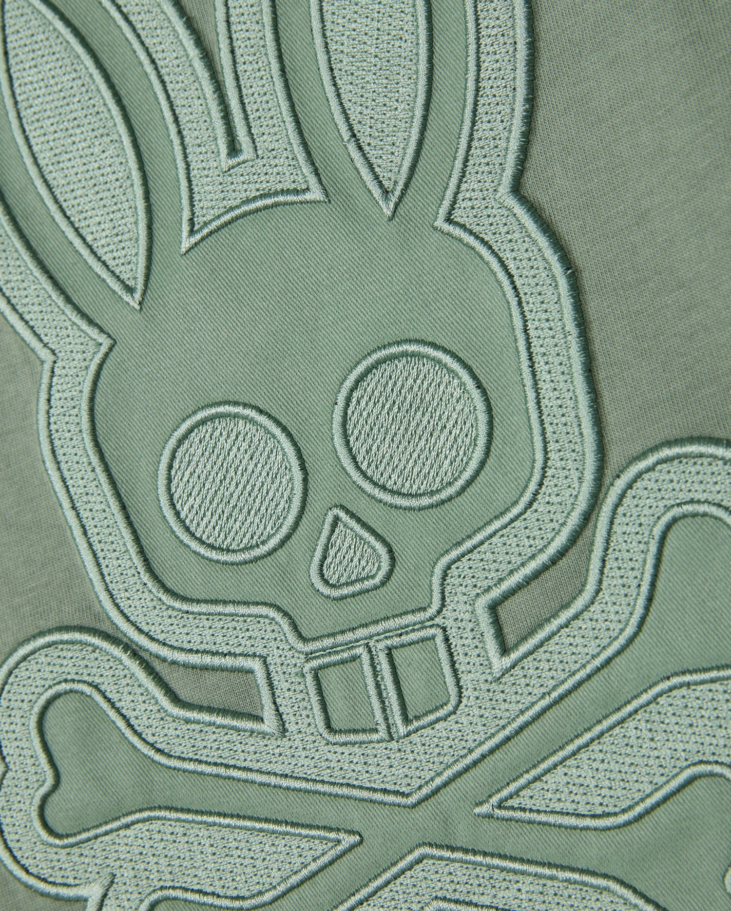 A close-up shot of the embroidered patch on the Pima cotton fabric background of the Psycho Bunny MENS MALTON GRAPHIC TEE - B6U795D200, showcasing a bunny skull and crossbones design in light green thread. The intricate stitching lends a three-dimensional feel to the design, echoing Peruvian-crafted artistry.