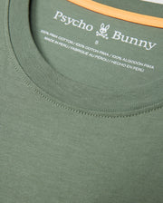 Close-up of a green MENS MALTON GRAPHIC TEE - B6U795D200 neckline with "Psycho Bunny" printed in white text. Below the brand name, the label states "100% Pima Cotton" and "Made in Peru" in both English and French. The size "6" is also indicated on this Peruvian-crafted label.