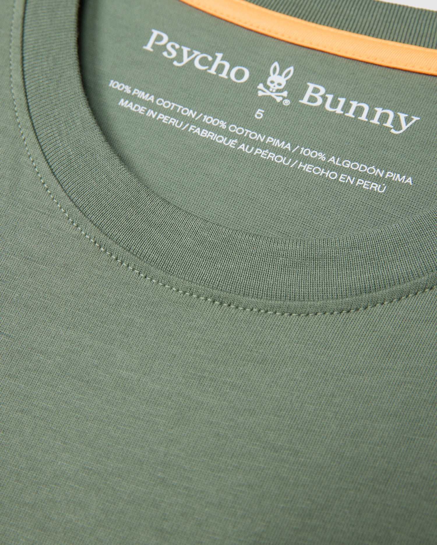 Close-up of a green MENS MALTON GRAPHIC TEE - B6U795D200 neckline with "Psycho Bunny" printed in white text. Below the brand name, the label states "100% Pima Cotton" and "Made in Peru" in both English and French. The size "6" is also indicated on this Peruvian-crafted label.