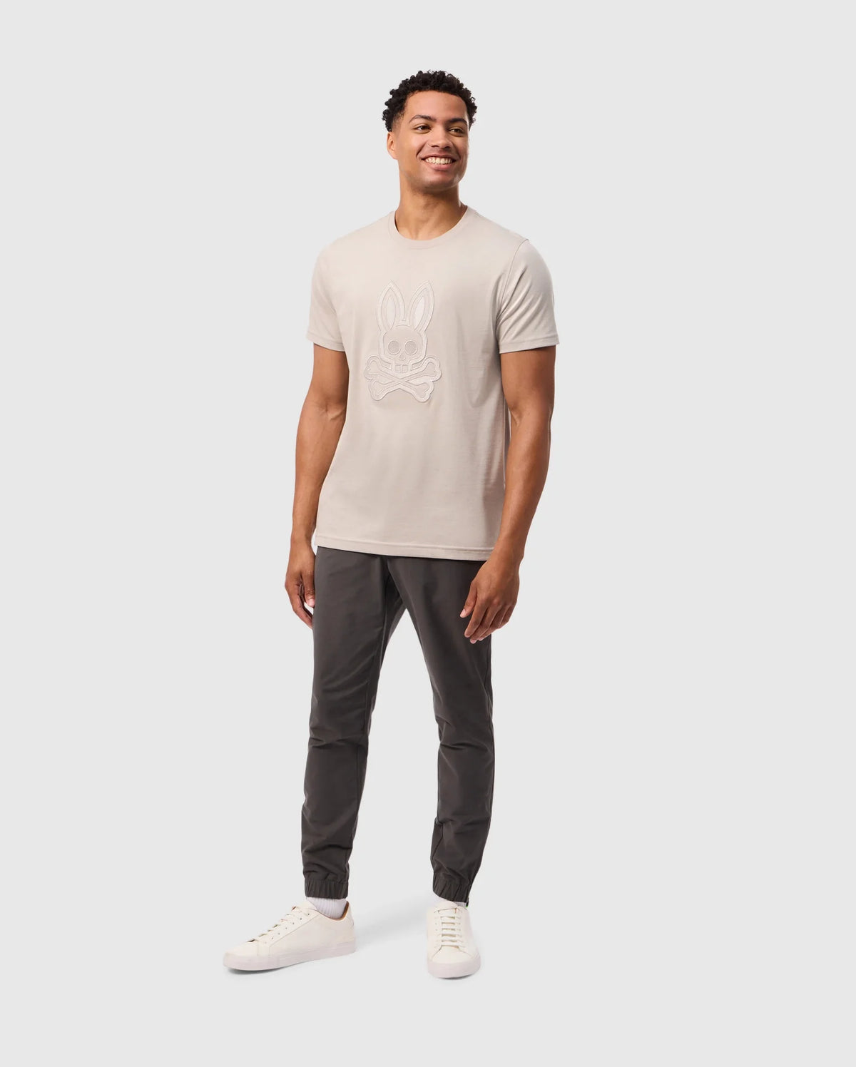 A smiling man stands against a plain white background, wearing a light-colored Psycho Bunny Mens Malton Graphic Tee (B6U795D200) made from soft Pima cotton with a cartoon rabbit on the front. Paired with dark jogger pants and white sneakers, his hands rest relaxed at his sides, showcasing effortless style and Peruvian craftsmanship.