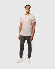 A smiling man stands against a plain white background, wearing a light-colored Psycho Bunny Mens Malton Graphic Tee (B6U795D200) made from soft Pima cotton with a cartoon rabbit on the front. Paired with dark jogger pants and white sneakers, his hands rest relaxed at his sides, showcasing effortless style and Peruvian craftsmanship.