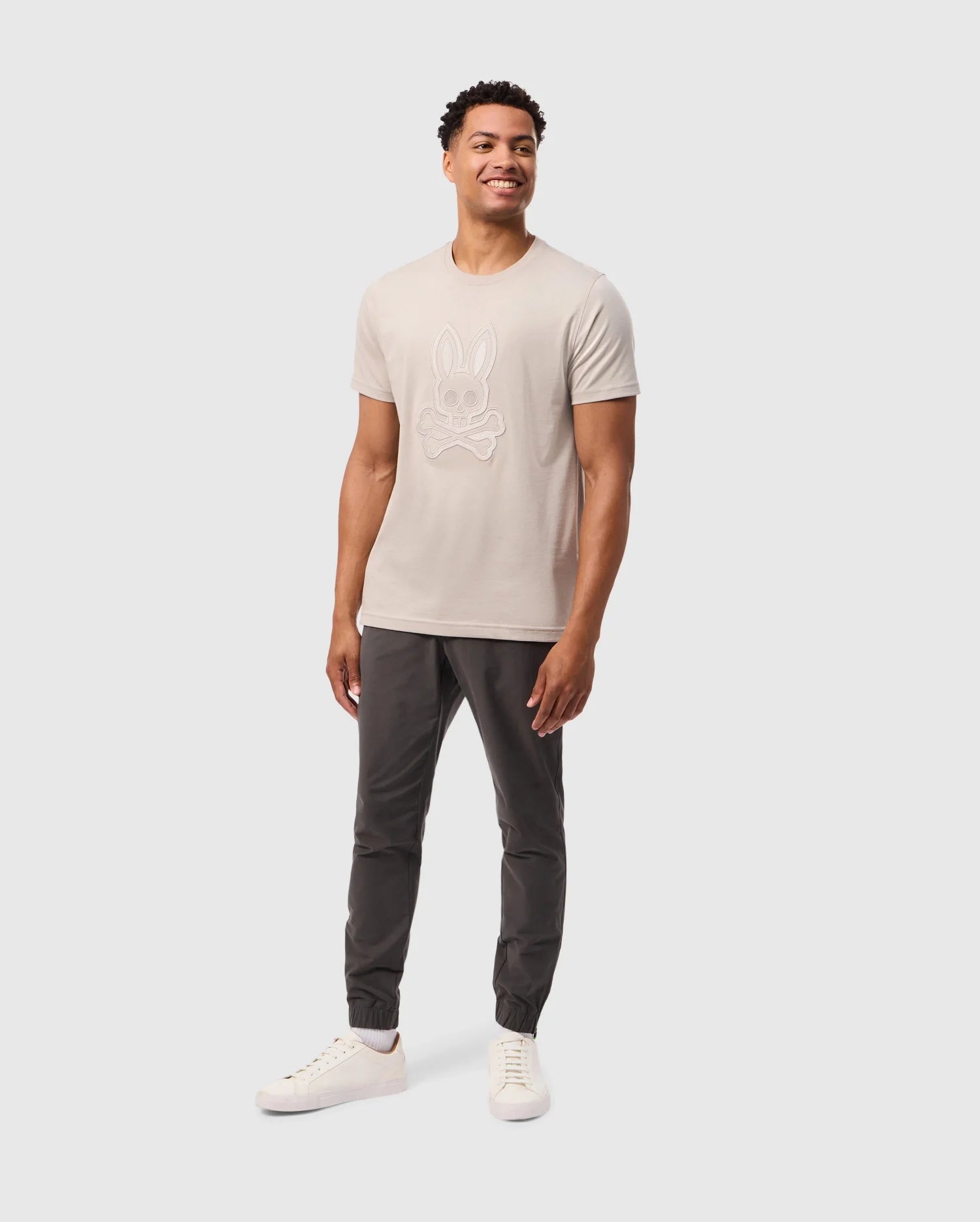 A smiling man stands against a plain white background, wearing a light-colored Psycho Bunny Mens Malton Graphic Tee (B6U795D200) made from soft Pima cotton with a cartoon rabbit on the front. Paired with dark jogger pants and white sneakers, his hands rest relaxed at his sides, showcasing effortless style and Peruvian craftsmanship.