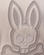 A close-up image showcasing an embroidered design of a stylized bunny head with elongated ears and a skull-like face, complete with crossbones below it, all on beige Pima cotton fabric. The design is outlined in white thread, demonstrating exquisite Peruvian craftsmanship. Product: MENS MALTON GRAPHIC TEE - B6U795D200 by Psycho Bunny.