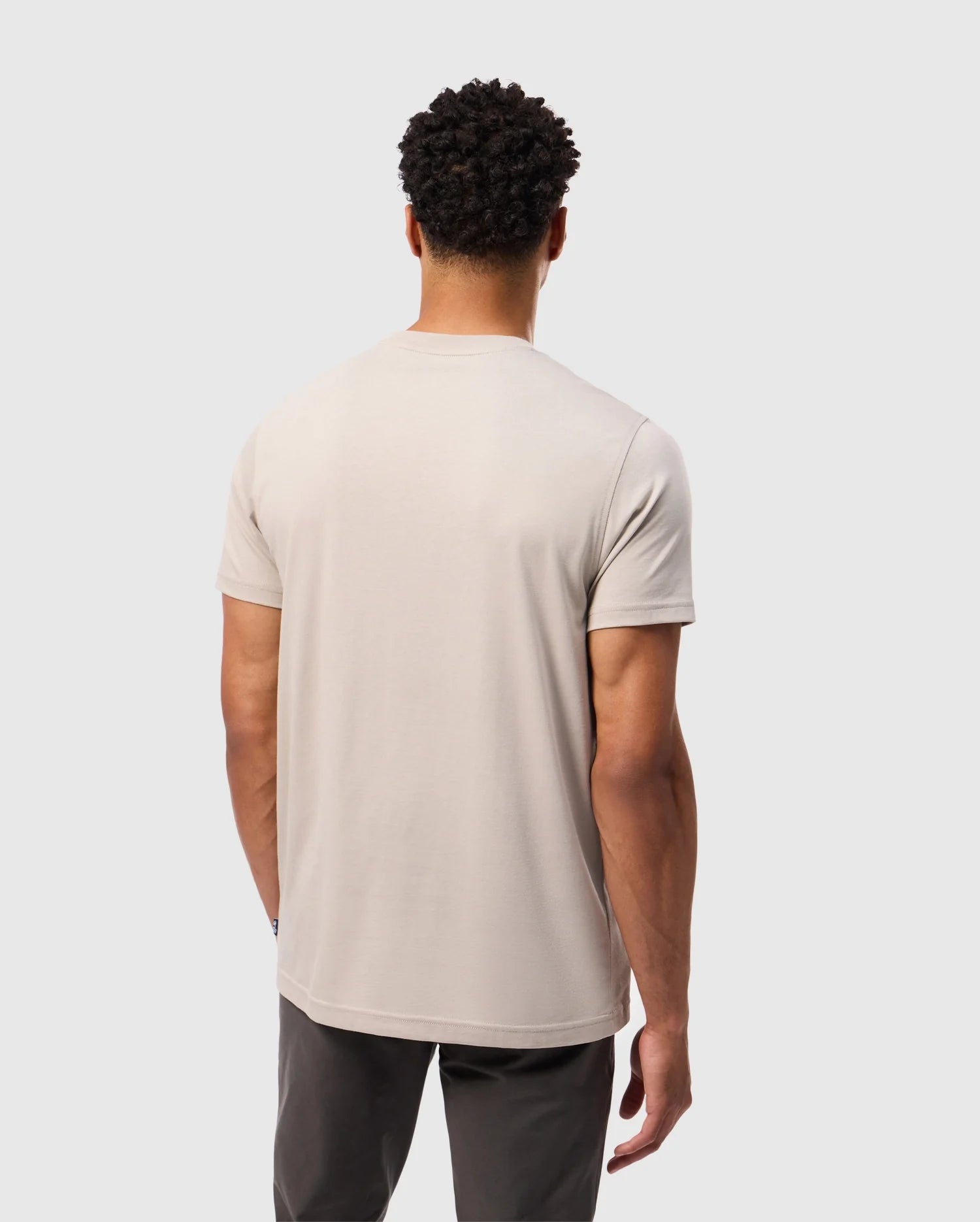 A man with short curly hair stands facing away, wearing a light beige Psycho Bunny MENS MALTON GRAPHIC TEE - B6U795D200 made from Pima cotton and dark pants. The background is a plain, light gray.