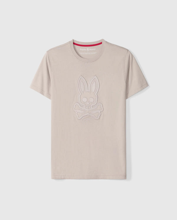 The Psycho Bunny Men's Malton Graphic Tee - B6U795D200 is a beige-colored T-shirt made from Pima cotton, featuring short sleeves and a round neck. It boasts an embossed design of a rabbit skull and crossbones. Highlighting Peruvian craftsmanship, the inner collar includes a red strip with white text. The T-shirt is showcased against a plain white background.