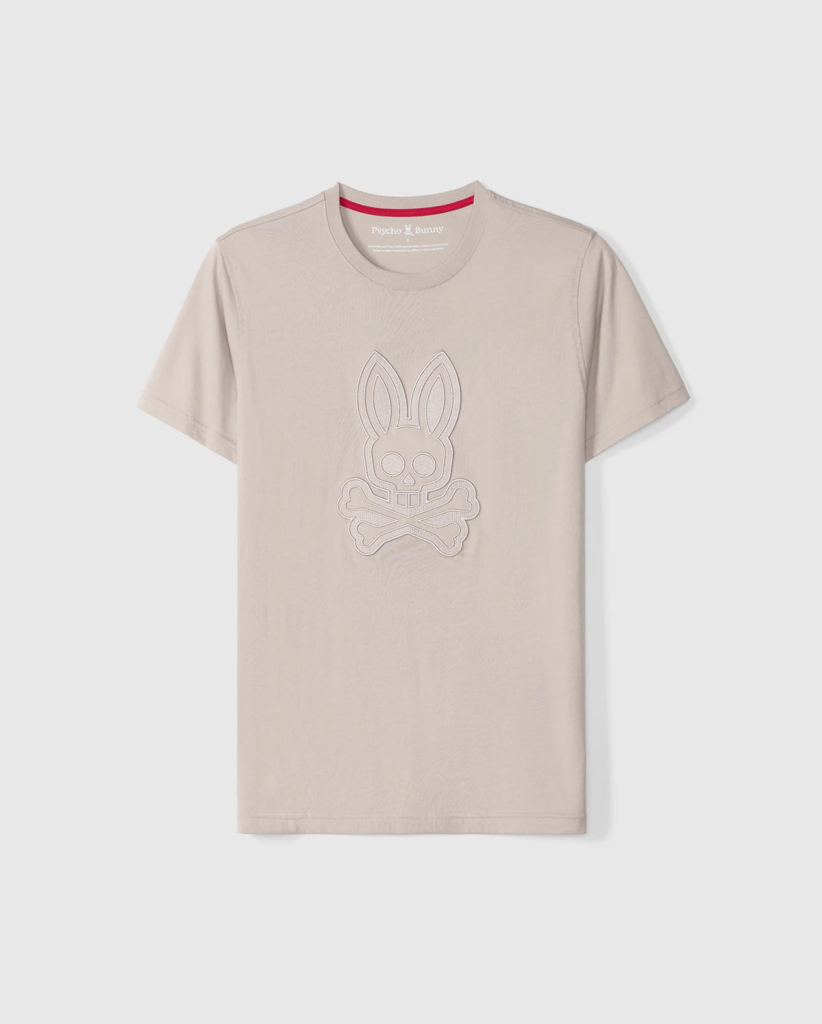 The Psycho Bunny Men's Malton Graphic Tee - B6U795D200 is a beige-colored T-shirt made from Pima cotton, featuring short sleeves and a round neck. It boasts an embossed design of a rabbit skull and crossbones. Highlighting Peruvian craftsmanship, the inner collar includes a red strip with white text. The T-shirt is showcased against a plain white background.