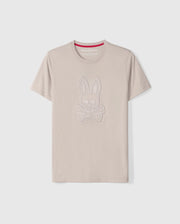 The Psycho Bunny Men's Malton Graphic Tee - B6U795D200 is a beige-colored T-shirt made from Pima cotton, featuring short sleeves and a round neck. It boasts an embossed design of a rabbit skull and crossbones. Highlighting Peruvian craftsmanship, the inner collar includes a red strip with white text. The T-shirt is showcased against a plain white background.
