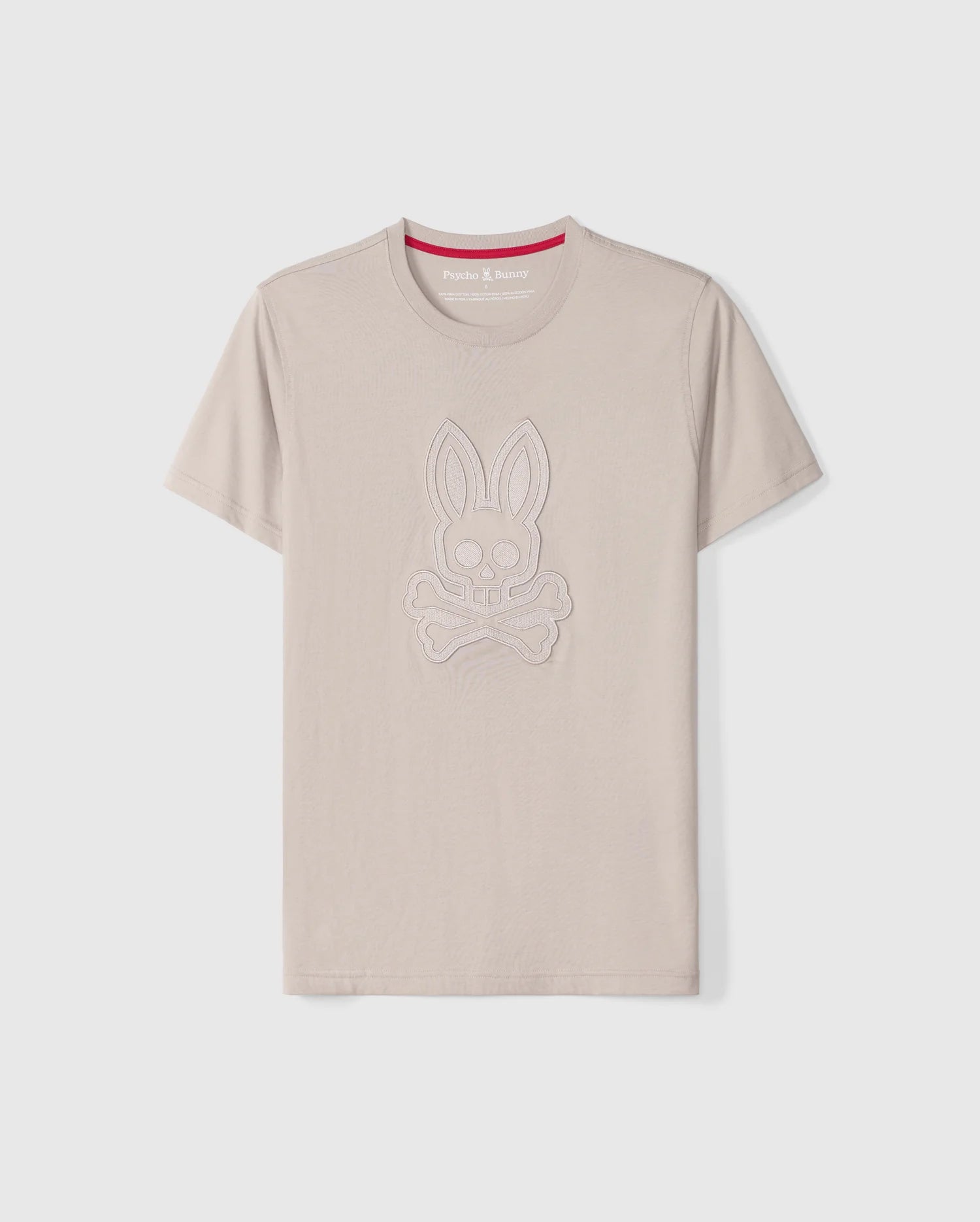The Psycho Bunny Men's Malton Graphic Tee - B6U795D200 is a beige-colored T-shirt made from Pima cotton, featuring short sleeves and a round neck. It boasts an embossed design of a rabbit skull and crossbones. Highlighting Peruvian craftsmanship, the inner collar includes a red strip with white text. The T-shirt is showcased against a plain white background.