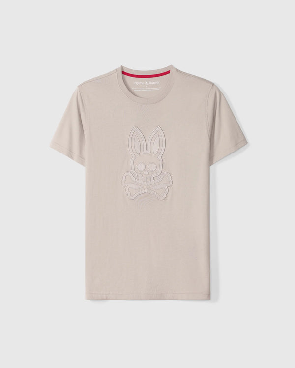 The Psycho Bunny Men's Malton Graphic Tee - B6U795D200 is a beige-colored T-shirt made from Pima cotton, featuring short sleeves and a round neck. It boasts an embossed design of a rabbit skull and crossbones. Highlighting Peruvian craftsmanship, the inner collar includes a red strip with white text. The T-shirt is showcased against a plain white background.