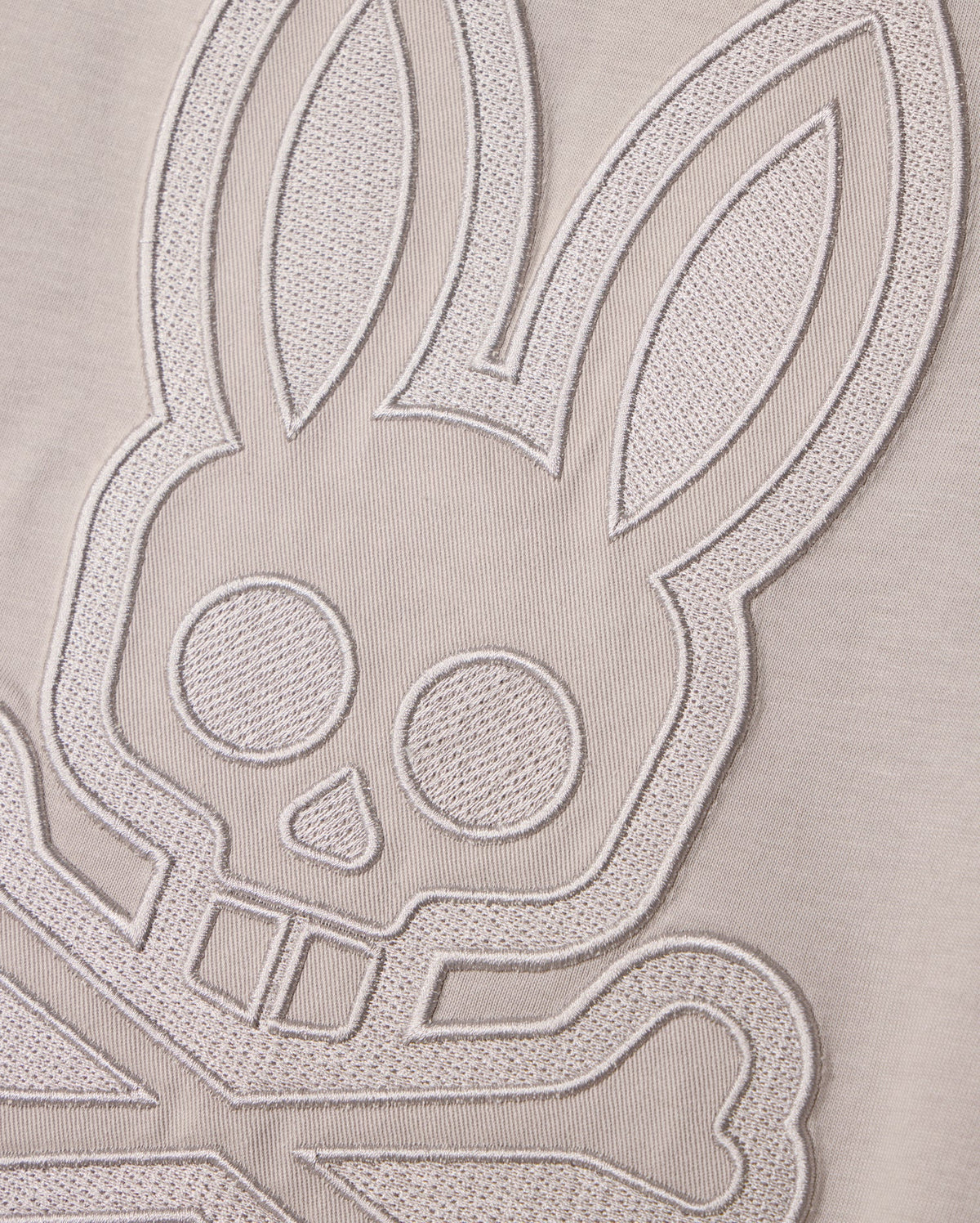 A close-up shot of textured embroidery on a MENS MALTON GRAPHIC TEE - B6U795D200 by Psycho Bunny, depicting a stylized bunny face with crossed bones underneath, reminiscent of a skull and crossbones design. The intricate embroidery is rendered in light, monochromatic thread on soft Pima cotton, showcasing exquisite Peruvian craftsmanship.