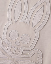 A close-up shot of textured embroidery on a MENS MALTON GRAPHIC TEE - B6U795D200 by Psycho Bunny, depicting a stylized bunny face with crossed bones underneath, reminiscent of a skull and crossbones design. The intricate embroidery is rendered in light, monochromatic thread on soft Pima cotton, showcasing exquisite Peruvian craftsmanship.