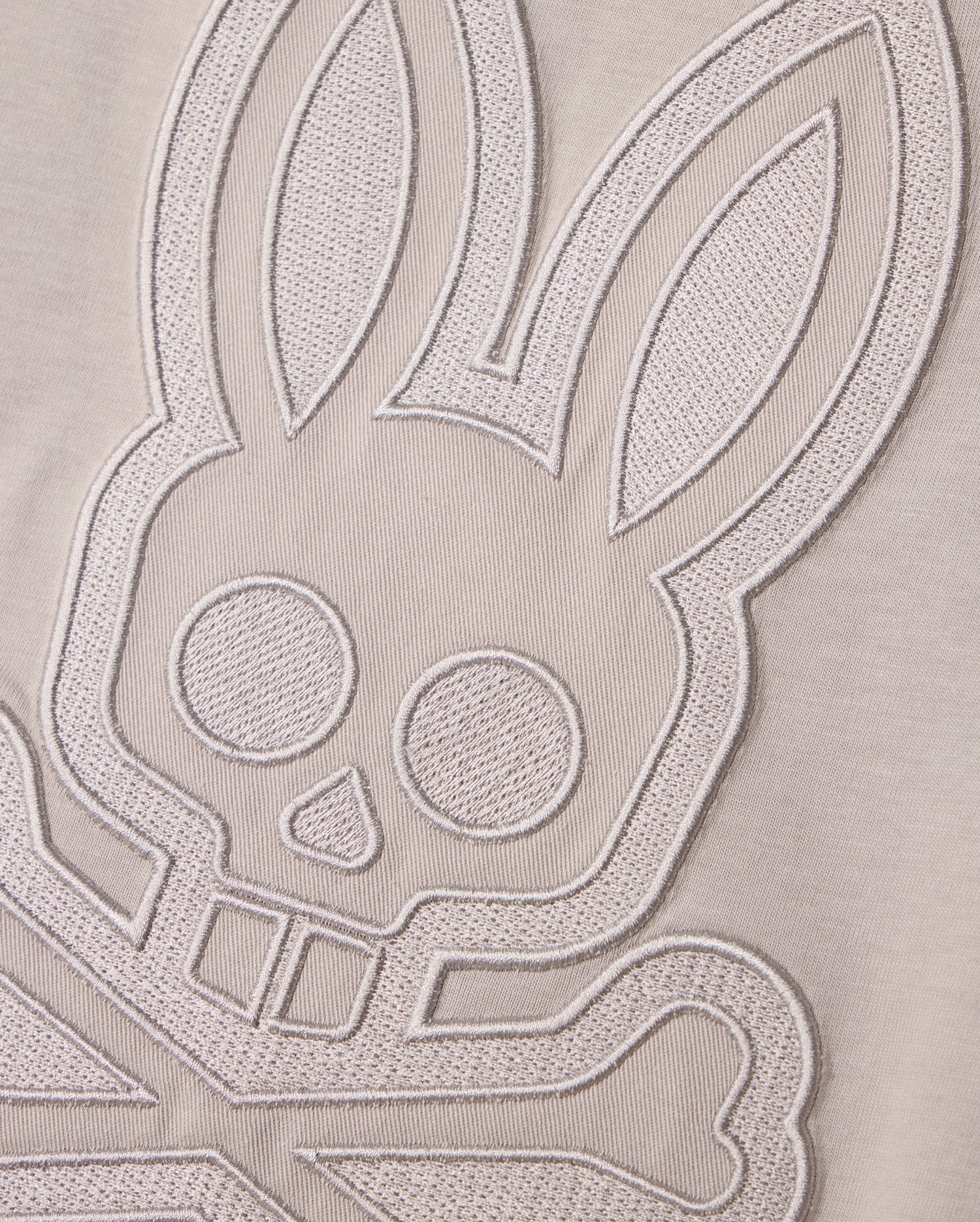 A close-up shot of textured embroidery on a MENS MALTON GRAPHIC TEE - B6U795D200 by Psycho Bunny, depicting a stylized bunny face with crossed bones underneath, reminiscent of a skull and crossbones design. The intricate embroidery is rendered in light, monochromatic thread on soft Pima cotton, showcasing exquisite Peruvian craftsmanship.