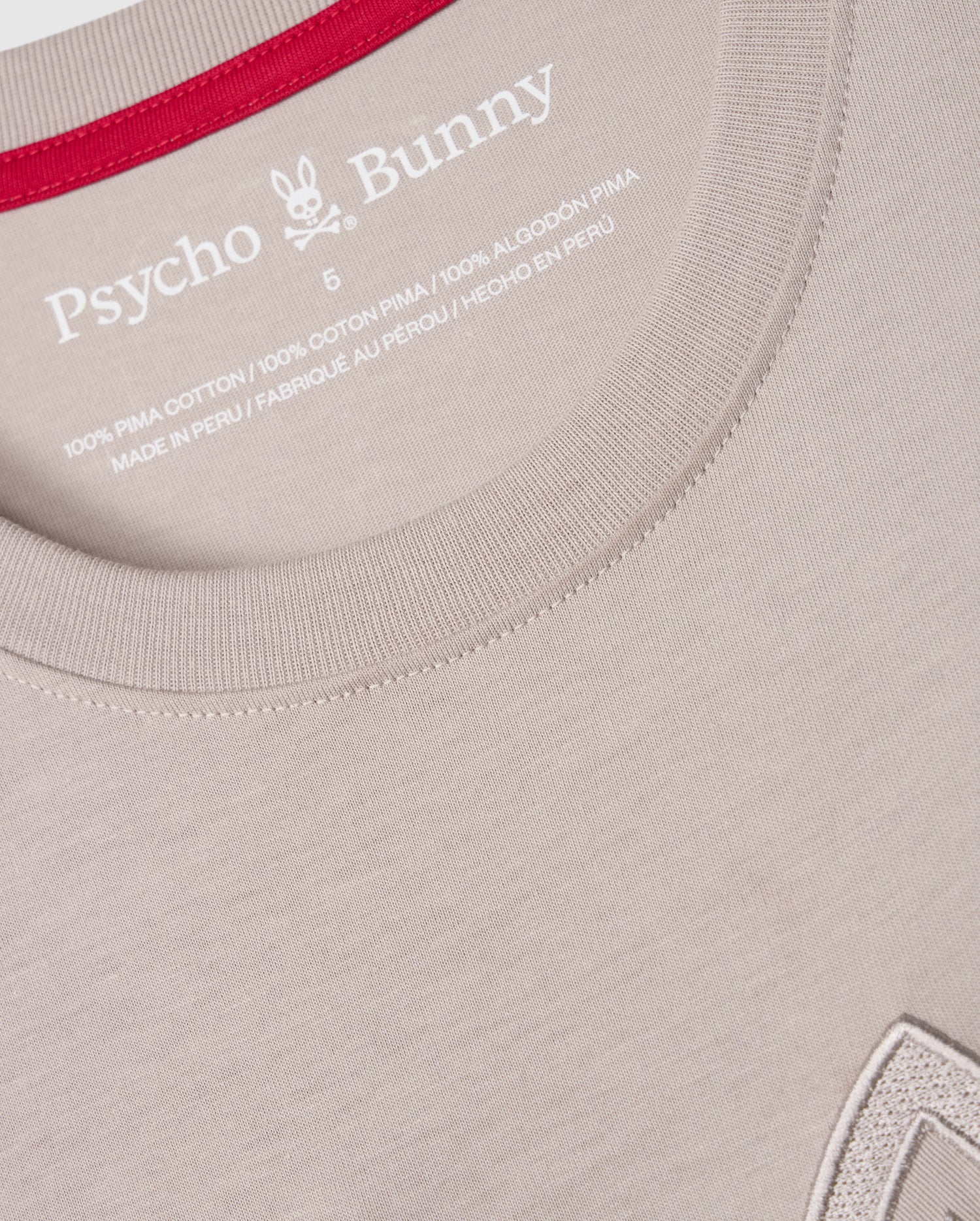 Close-up image of a light beige Psycho Bunny MENS MALTON GRAPHIC TEE - B6U795D200 featuring the brand logo and text printed in white on the inside of the collar. The crew neck has a red inner lining, and a small portion of a graphic can be seen at the bottom right of the image, showcasing Peruvian craftsmanship.