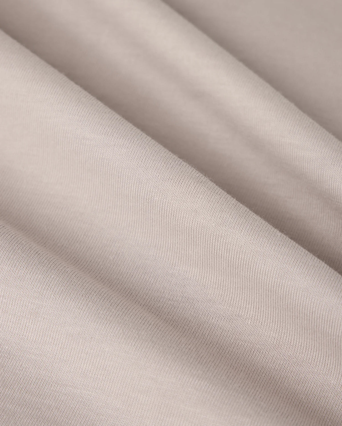 A close-up image of the MENS MALTON GRAPHIC TEE by Psycho Bunny, showcasing light beige fabric with a smooth texture and subtle diagonal folds, reminiscent of the fine weave found in Pima cotton. The material appears soft and finely woven, creating gentle shadows and highlights across its surface.