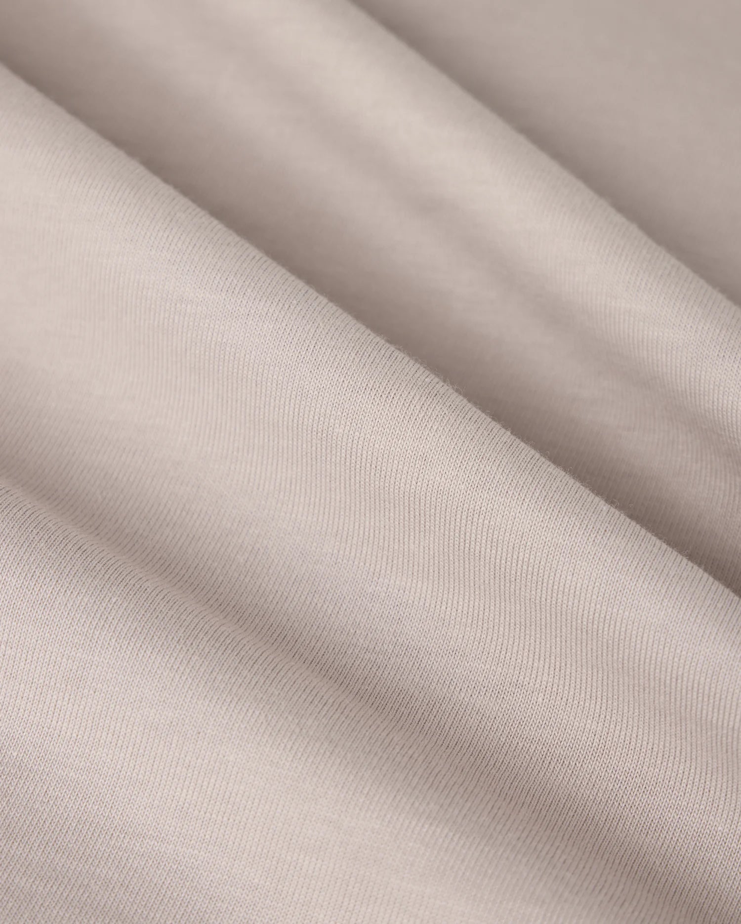A close-up image of the MENS MALTON GRAPHIC TEE by Psycho Bunny, showcasing light beige fabric with a smooth texture and subtle diagonal folds, reminiscent of the fine weave found in Pima cotton. The material appears soft and finely woven, creating gentle shadows and highlights across its surface.