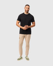 A man wearing a black Psycho Bunny MENS CAMPO POCKET TEE - B6U801D200 and beige pants stands against a light gray background. He has short hair and a beard, with his hands clasped together, completing his look with green sneakers.