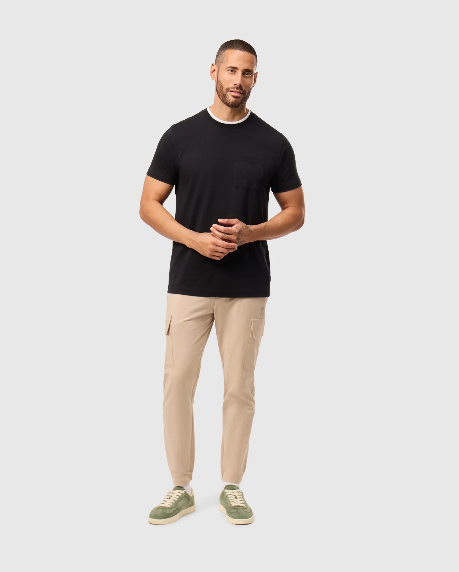 A man wearing a black Psycho Bunny MENS CAMPO POCKET TEE - B6U801D200 and beige pants stands against a light gray background. He has short hair and a beard, with his hands clasped together, completing his look with green sneakers.