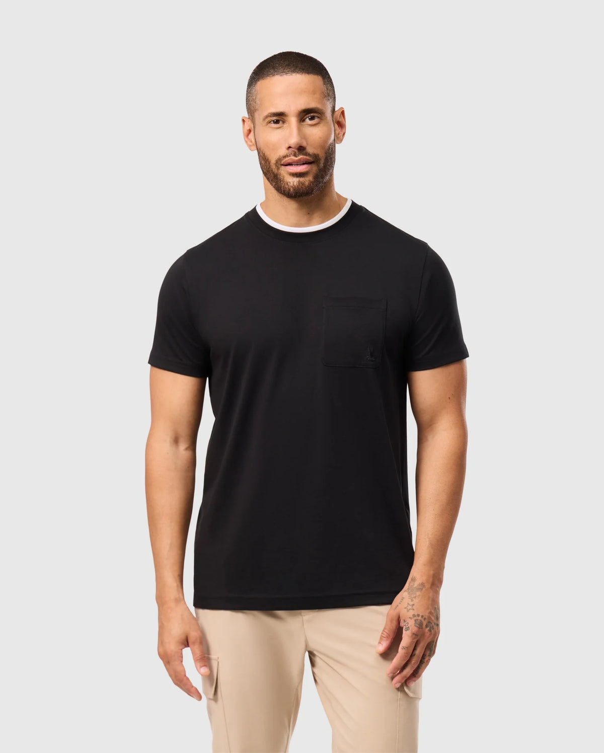 A man with short hair and a beard is wearing a black Psycho Bunny MENS CAMPO POCKET TEE - B6U801D200 and beige pants. He has tattoos on his left hand and stands against a plain light gray background.