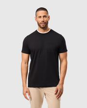 A man with short hair and a beard is wearing a black Psycho Bunny MENS CAMPO POCKET TEE - B6U801D200 and beige pants. He has tattoos on his left hand and stands against a plain light gray background.
