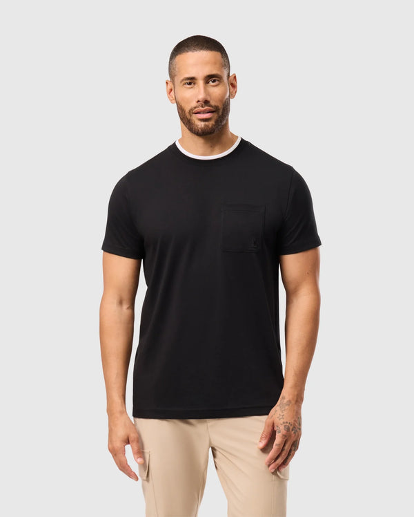 A man with short hair and a beard is wearing a black Psycho Bunny MENS CAMPO POCKET TEE - B6U801D200 and beige pants. He has tattoos on his left hand and stands against a plain light gray background.