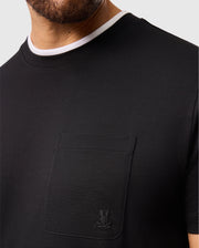 A person wearing the Psycho Bunny MENS CAMPO POCKET TEE - B6U801D200 in black, featuring a white double collar with a small embroidered logo. The soft pima cotton material is emphasized in the T-shirt's design. The person's face is partially visible, displaying short facial hair.