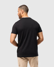 A person with short hair wears the Psycho Bunny MENS CAMPO POCKET TEE - B6U801D200 in black, crafted from pima cotton, paired with beige pants, and stands facing away from the camera against a plain gray background.
