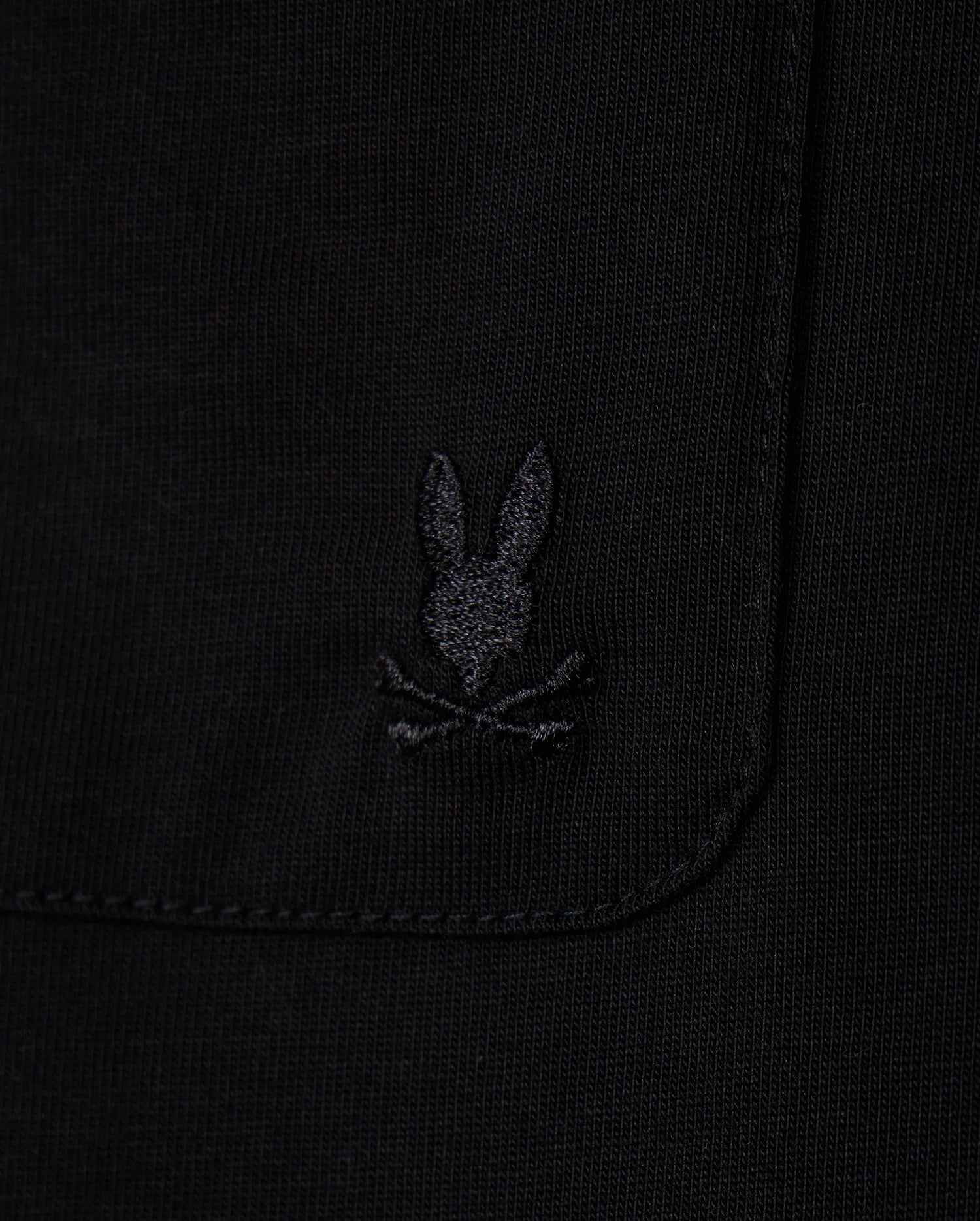 Close-up of the MENS CAMPO POCKET TEE by Psycho Bunny, a black pocket tee made from premium pima cotton, showcasing a subtle embroidered logo with a stylized rabbit's head above crossed bones.