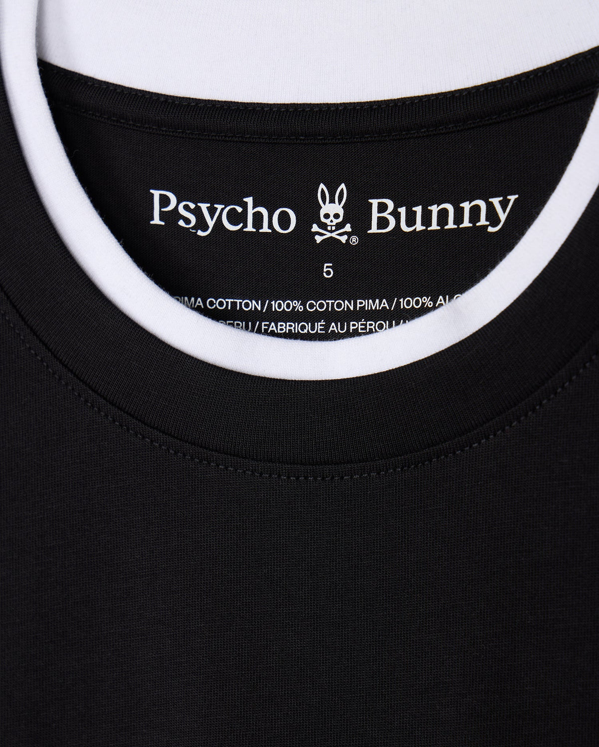 Close-up of a black and white double collar T-shirt, identified as the "MENS CAMPO POCKET TEE - B6U801D200," featuring a label with "Psycho Bunny" text and a bunny logo. The label states that it is made of 100% Pima cotton material and manufactured in Peru.