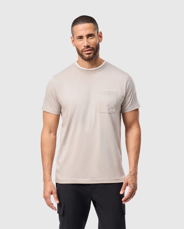 A man with short hair and a beard wears a Psycho Bunny MENS CAMPO POCKET TEE in light gray, made from soft pima cotton. The tee pairs effortlessly with his black pants as he stands against a plain white background.