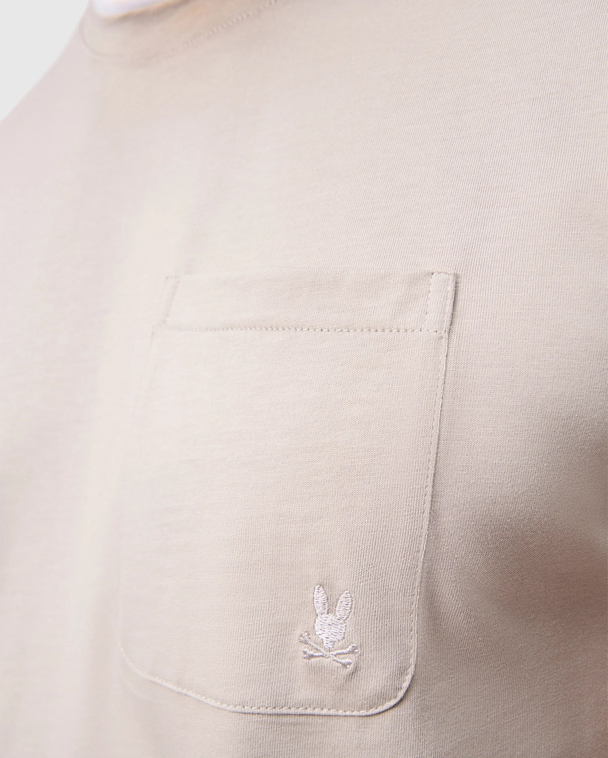 Close-up of the MENS CAMPO POCKET TEE by Psycho Bunny, featuring a beige pima cotton fabric with a chest pocket adorned with a white embroidered bunny and crossbones.