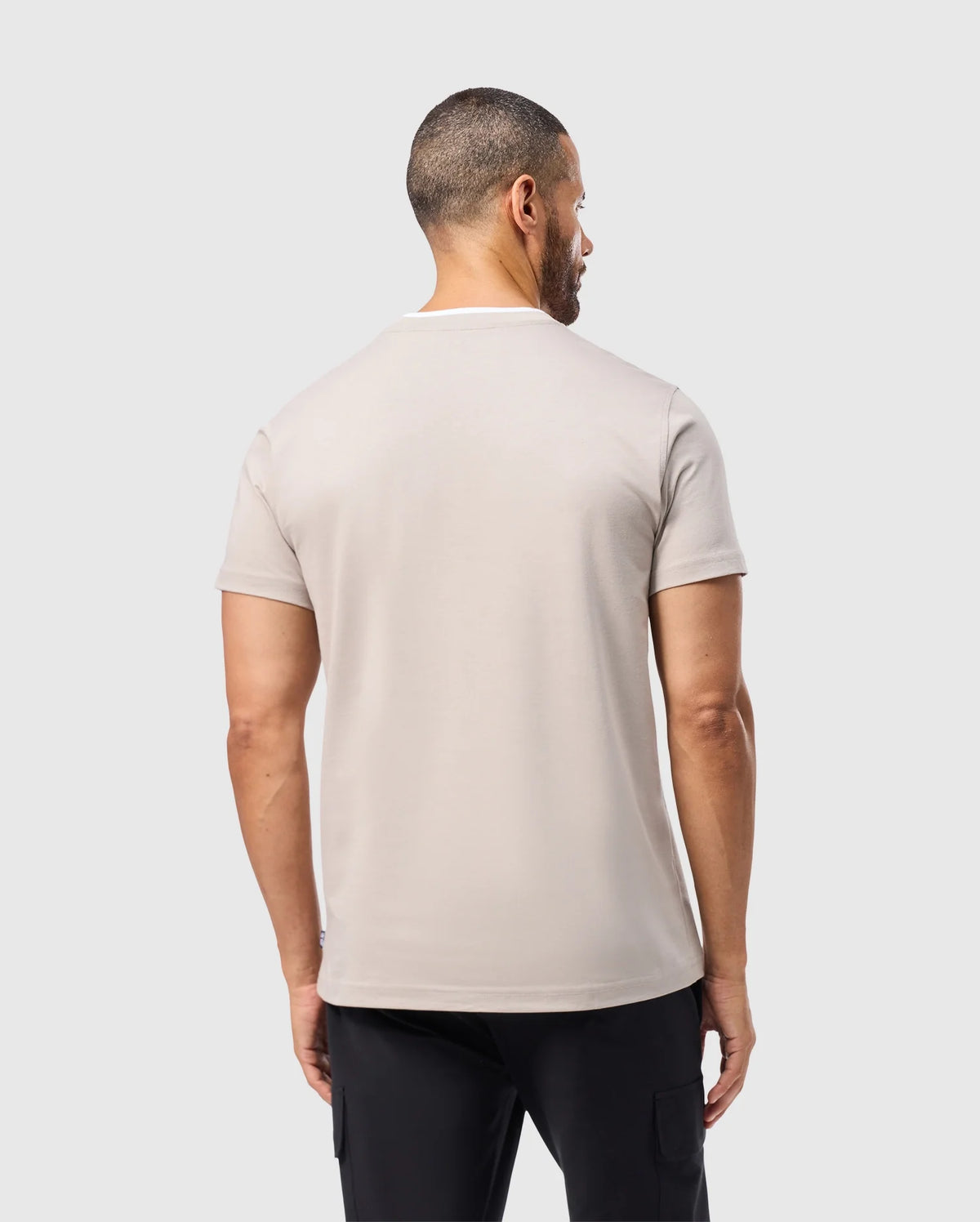 A man wearing the Psycho Bunny MENS CAMPO POCKET TEE in light beige, made from soft pima cotton, and black pants stands with his back to the camera against a plain gray background.