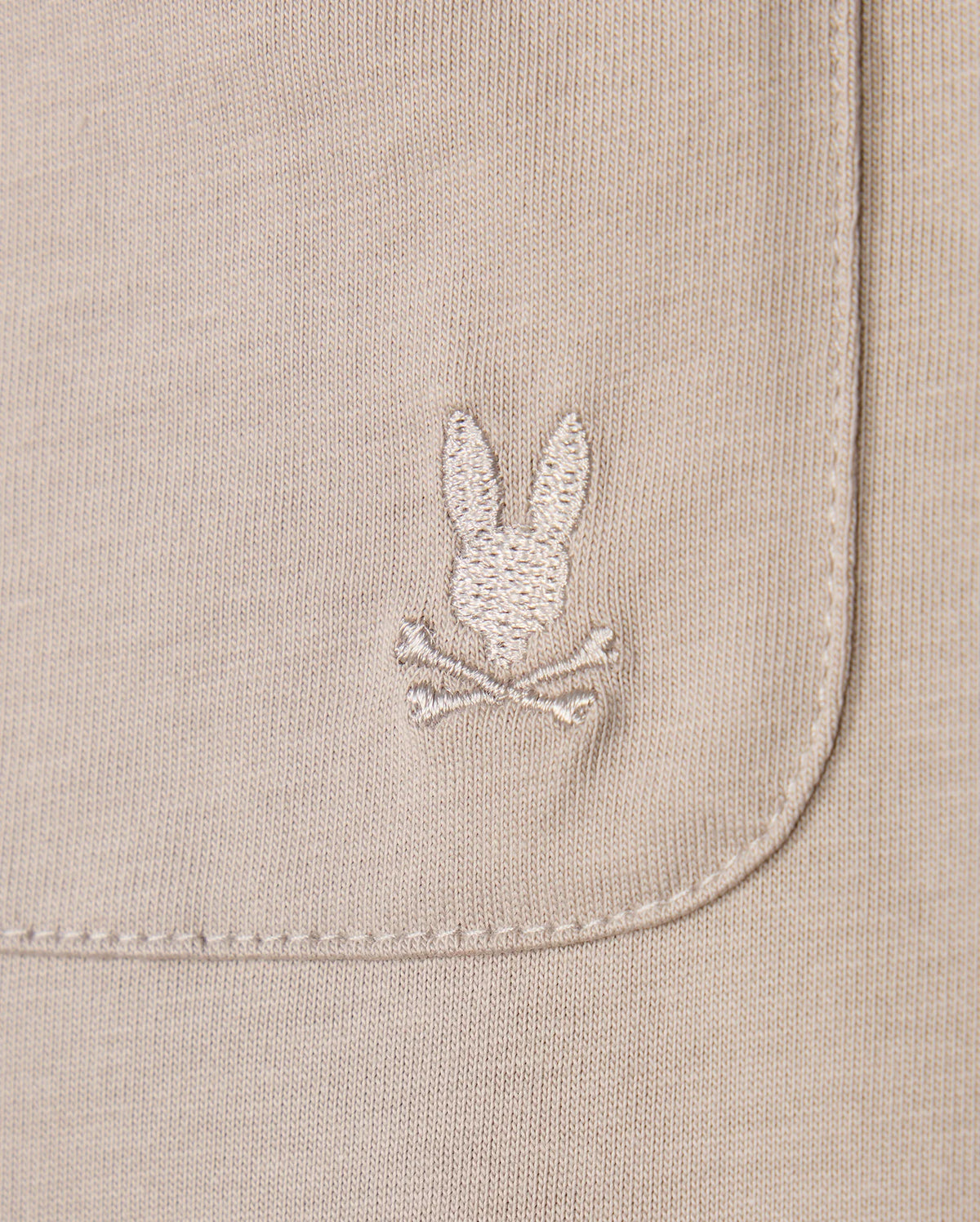 Close-up of the Psycho Bunny MENS CAMPO POCKET TEE - B6U801D200 in beige pima cotton, showcasing an embroidered rabbit's head and crossbones on the pocket. The texture and detailed stitching are clearly visible.