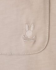 Close-up of the Psycho Bunny MENS CAMPO POCKET TEE - B6U801D200 in beige pima cotton, showcasing an embroidered rabbit's head and crossbones on the pocket. The texture and detailed stitching are clearly visible.