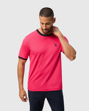 A man in a Psycho Bunny MENS EDMONDS SPORT TEE - B6U857D200 stands against a light gray background, sporting a bright pink tee with black trim on the collar and sleeves, and a small black emblem. Made from antimicrobial fabric, he has short hair and touches his neck while looking down.
