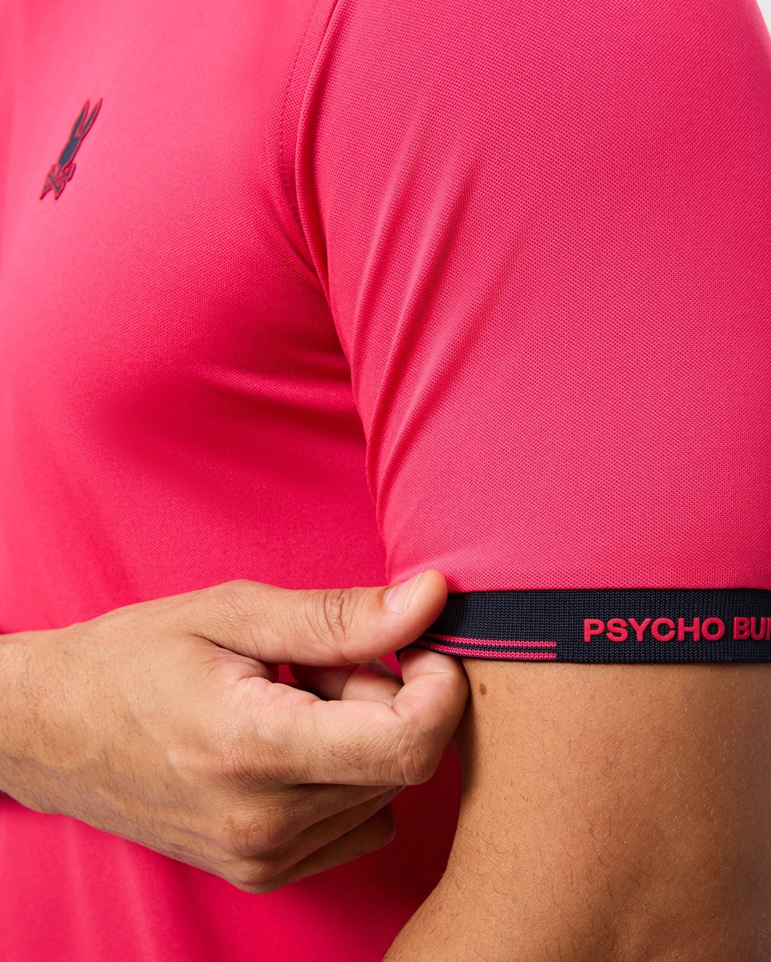 A person wears the MENS EDMONDS SPORT TEE by Psycho Bunny, featuring a small logo on the chest. The pink short-sleeve tee is made from antimicrobial fabric. They hold a sleeve edge with a black band reading "PSYCHO BUN" in red text.