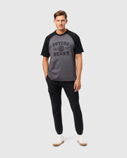 A man confidently wears a Psycho Bunny MENS LONDON RAGLAN GRAPHIC TEE - B6U896D200 made from premium Pima cotton in gray and black. He pairs it with black pants, white sneakers, and stands with his right hand in his pocket against a plain white background.