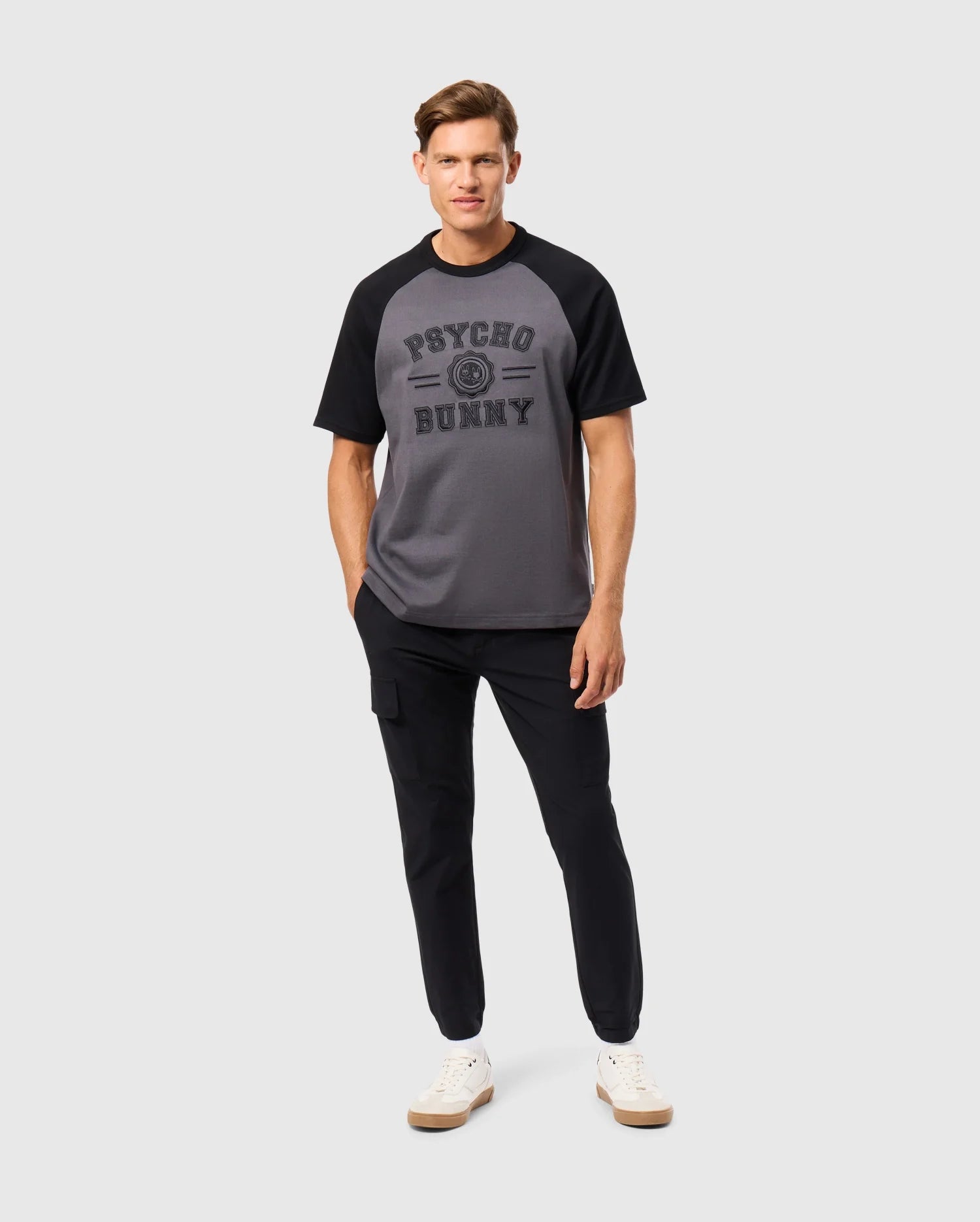 A man confidently wears a Psycho Bunny MENS LONDON RAGLAN GRAPHIC TEE - B6U896D200 made from premium Pima cotton in gray and black. He pairs it with black pants, white sneakers, and stands with his right hand in his pocket against a plain white background.