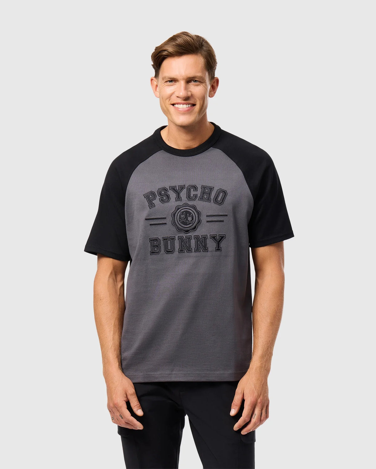 A smiling man stands against a plain background, wearing the MENS LONDON RAGLAN GRAPHIC TEE (B6U896D200) from Psycho Bunny, featuring gray and black short sleeves crafted from soft Pima cotton.