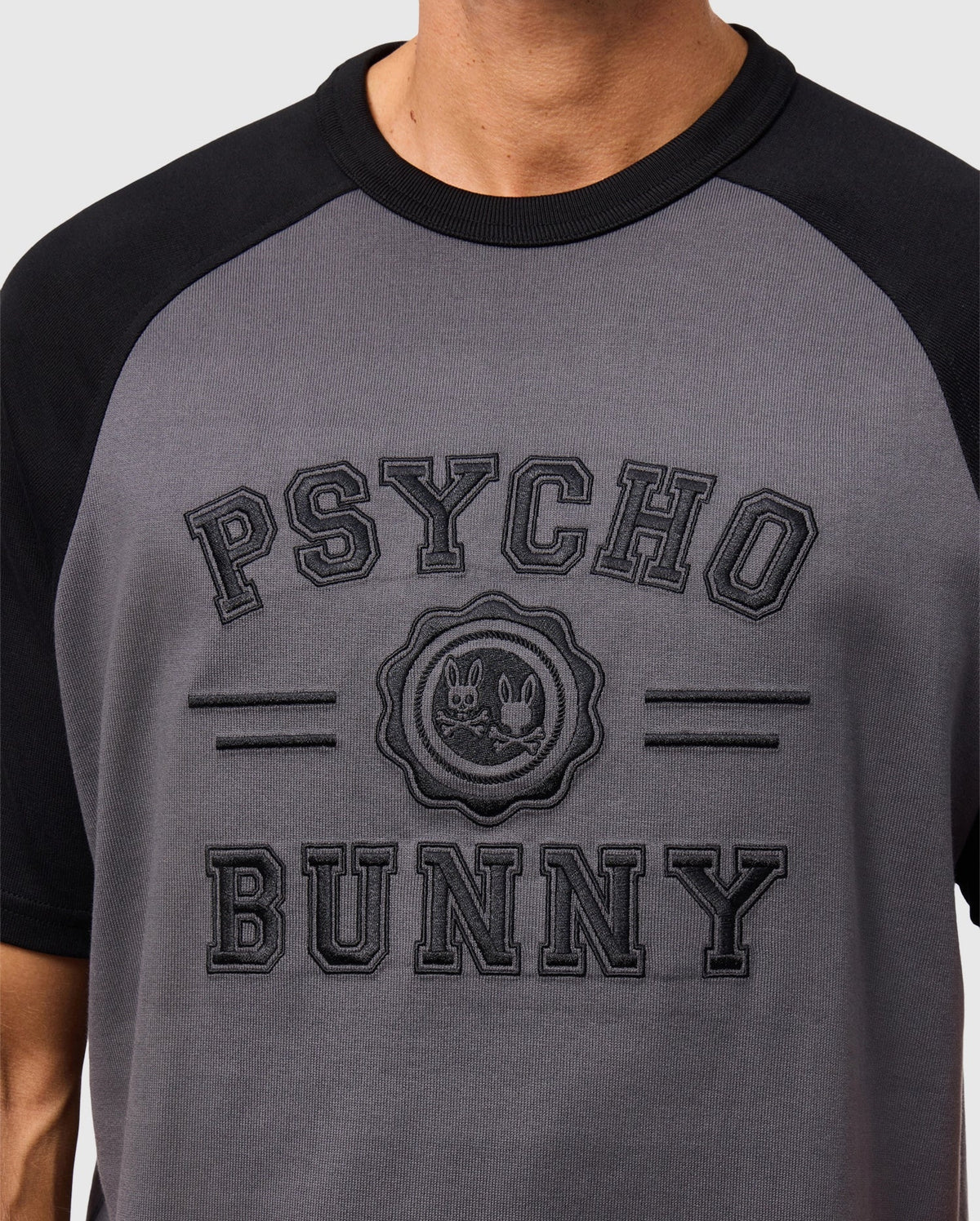 A person is wearing the Psycho Bunny Men's London Raglan Graphic Tee (B6U896D200) in gray and black. The heavyweight jersey tee, made from premium Pima cotton, features a bold "Psycho Bunny" logo with a bunny and skull motif.