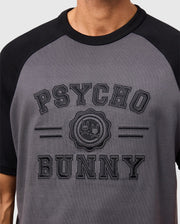 A person is wearing the Psycho Bunny Men's London Raglan Graphic Tee (B6U896D200) in gray and black. The heavyweight jersey tee, made from premium Pima cotton, features a bold "Psycho Bunny" logo with a bunny and skull motif.