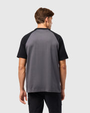 A brown-haired man faces away from the camera, wearing Psycho Bunny's MENS LONDON RAGLAN GRAPHIC TEE (B6U896D200) in gray and black. Crafted from soft Pima cotton, the tee is elegantly simple against a plain, light background.