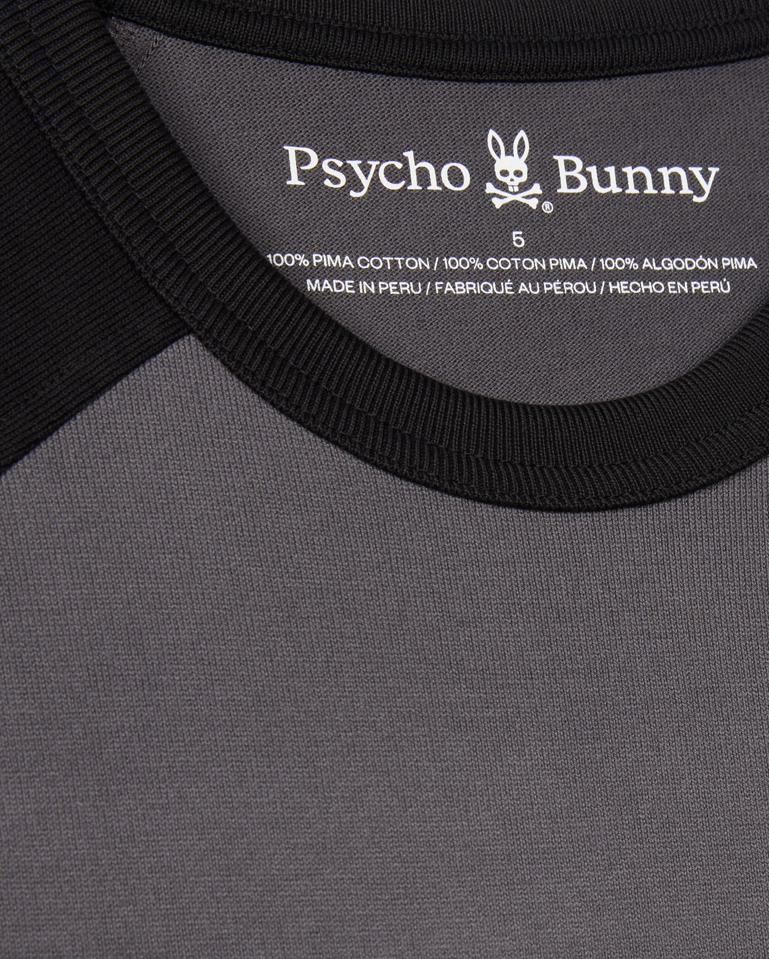 Close-up of the black and gray MENS LONDON RAGLAN GRAPHIC TEE - B6U896D200 by Psycho Bunny. It features a label on the inside neckline, is a men's graphic tee made of 100% Pima cotton, and is manufactured in Peru.