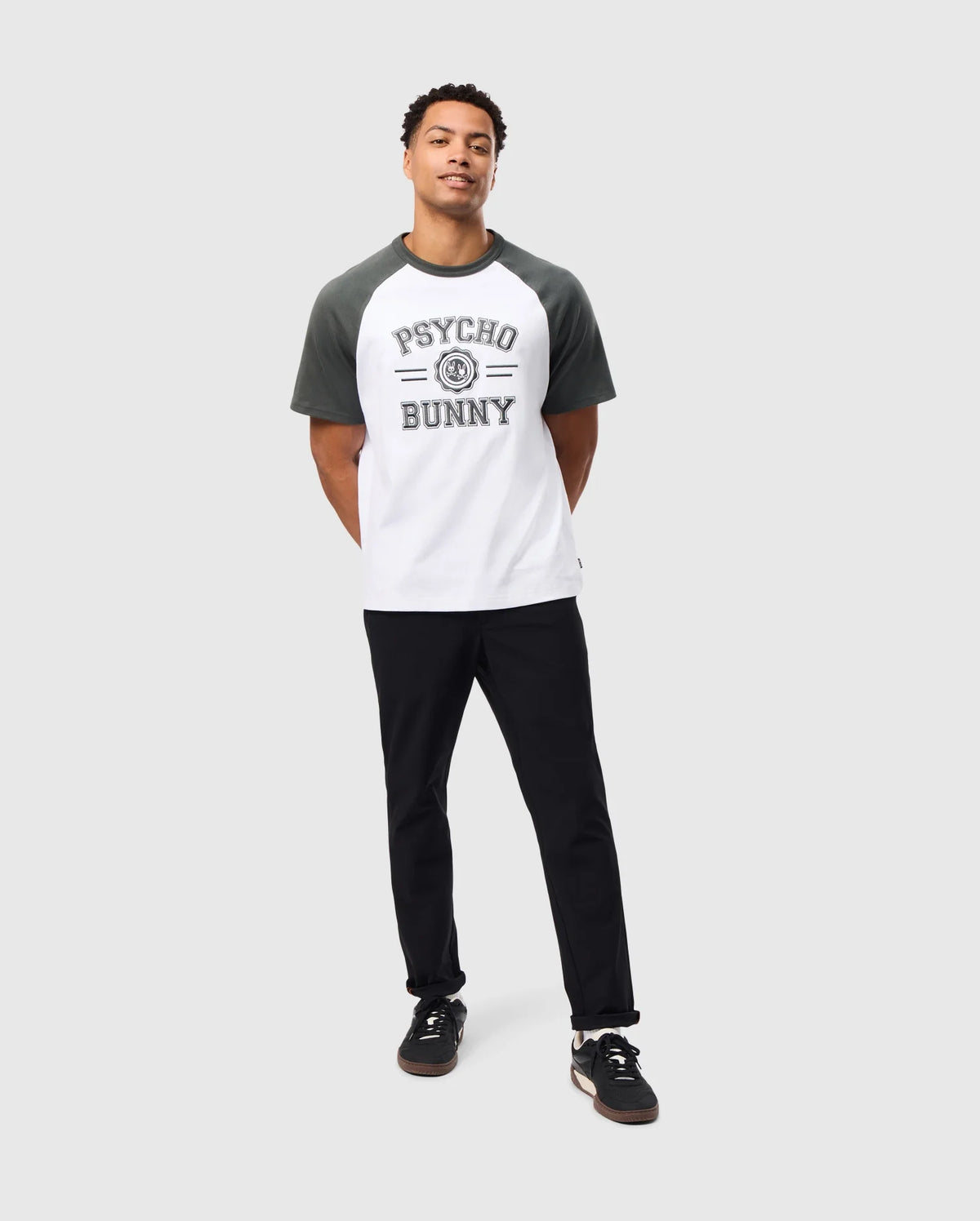 A man in the MENS LONDON RAGLAN GRAPHIC TEE - B6U896D200 by Psycho Bunny, designed in a heavy-weight black-and-white graphic style, stands against a light gray background. His outfit includes black pants and sneakers with white soles. Looking calmly upward, he exudes casual confidence with his hands behind his back.