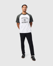 A man in the MENS LONDON RAGLAN GRAPHIC TEE - B6U896D200 by Psycho Bunny, designed in a heavy-weight black-and-white graphic style, stands against a light gray background. His outfit includes black pants and sneakers with white soles. Looking calmly upward, he exudes casual confidence with his hands behind his back.
