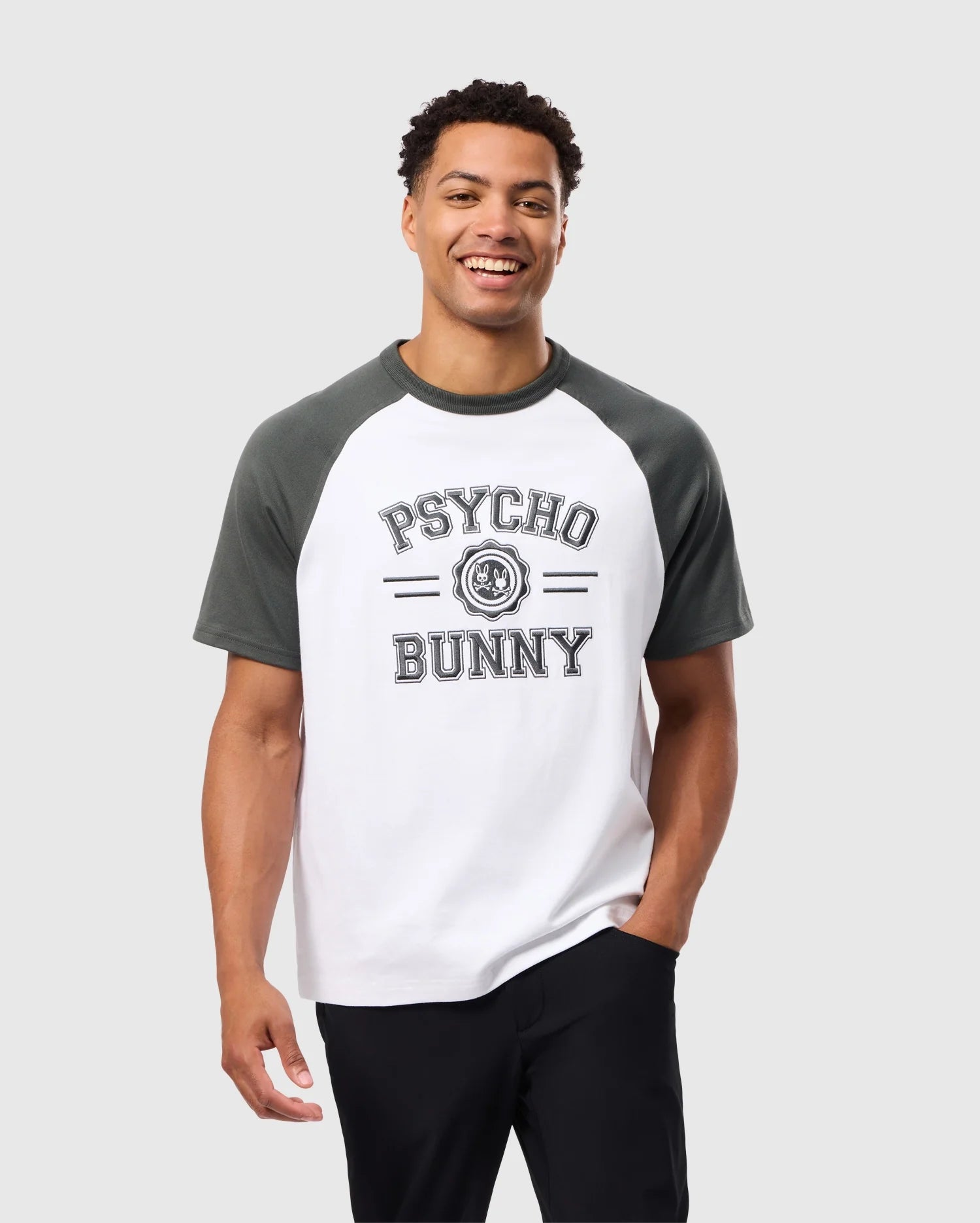 A smiling man stands against a simple backdrop, wearing the Psycho Bunny MENS LONDON RAGLAN GRAPHIC TEE - B6U896D200, crafted from soft Pima cotton. The white and gray raglan sleeves harmonize with the "Psycho Bunny" name and bunny logo on the front, perfectly paired with black pants for a varsity graphic look.
